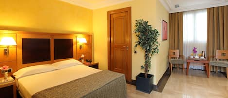 Habitación de hotel con una cama grande, dos lámparas de noche, una puerta de madera, una planta en maceta, un área de comedor con sillas y una mesa dispuesta con una tetera y tazas.