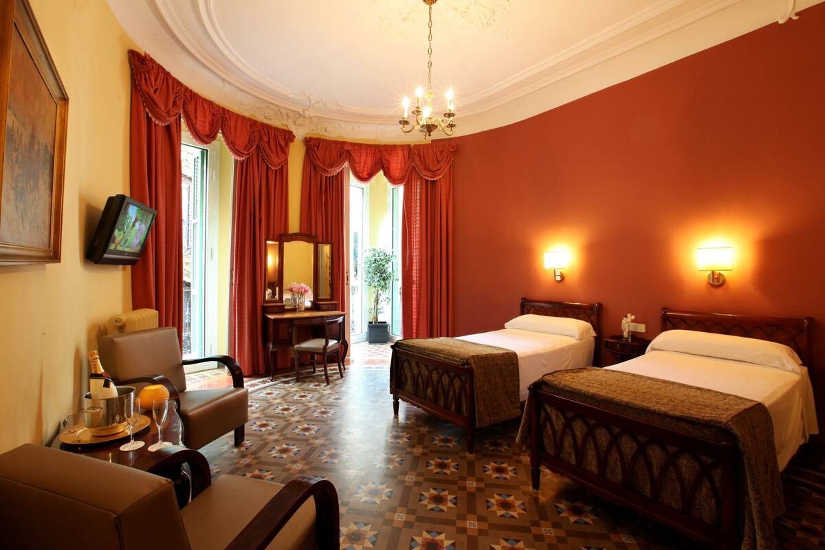 A hotel room with two beds, a desk with a chair, a television, and a chandelier.