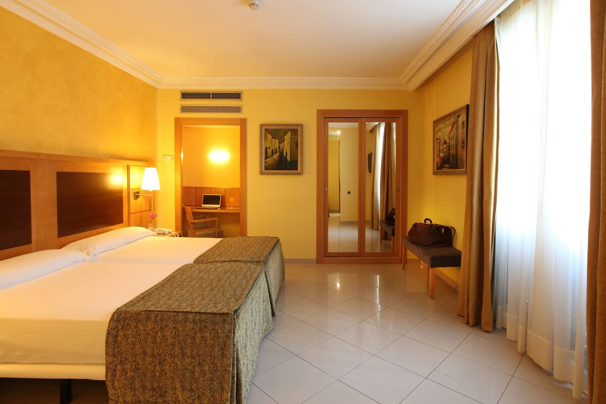 A hotel room with two beds, a desk, a chair, a lamp, a painting, and a door leading to another room.