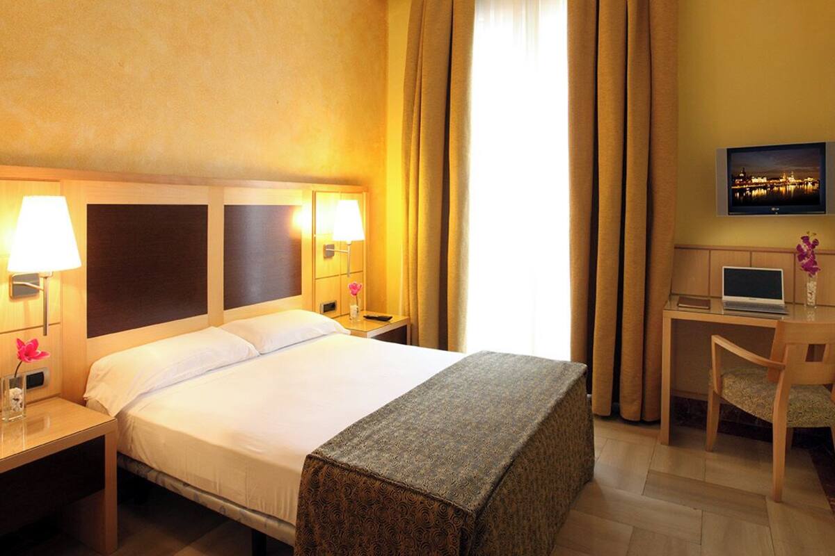 A hotel room with a bed, bedside lamps, a desk with a chair, a television, and a picture on the wall.