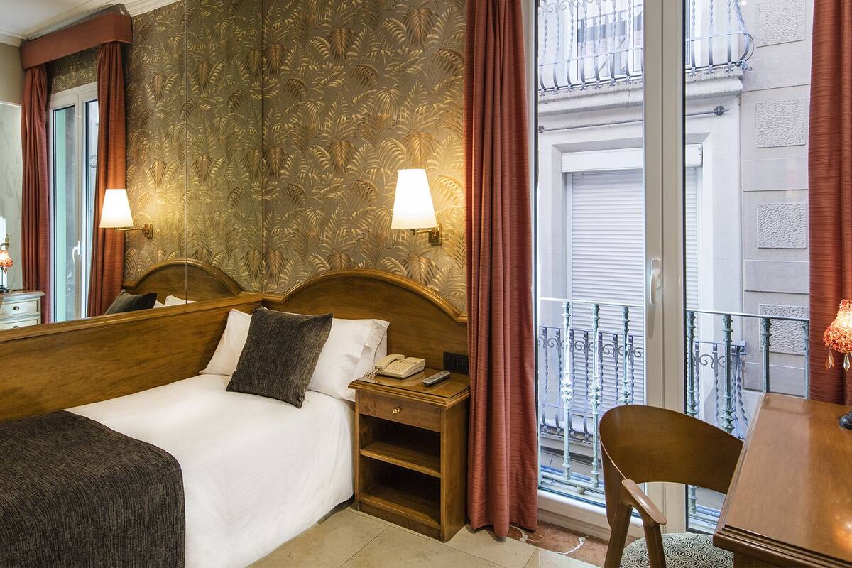 A hotel room with a large bed, two bedside lamps, a nightstand with a phone, and a view of the city through a balcony with metal railings.