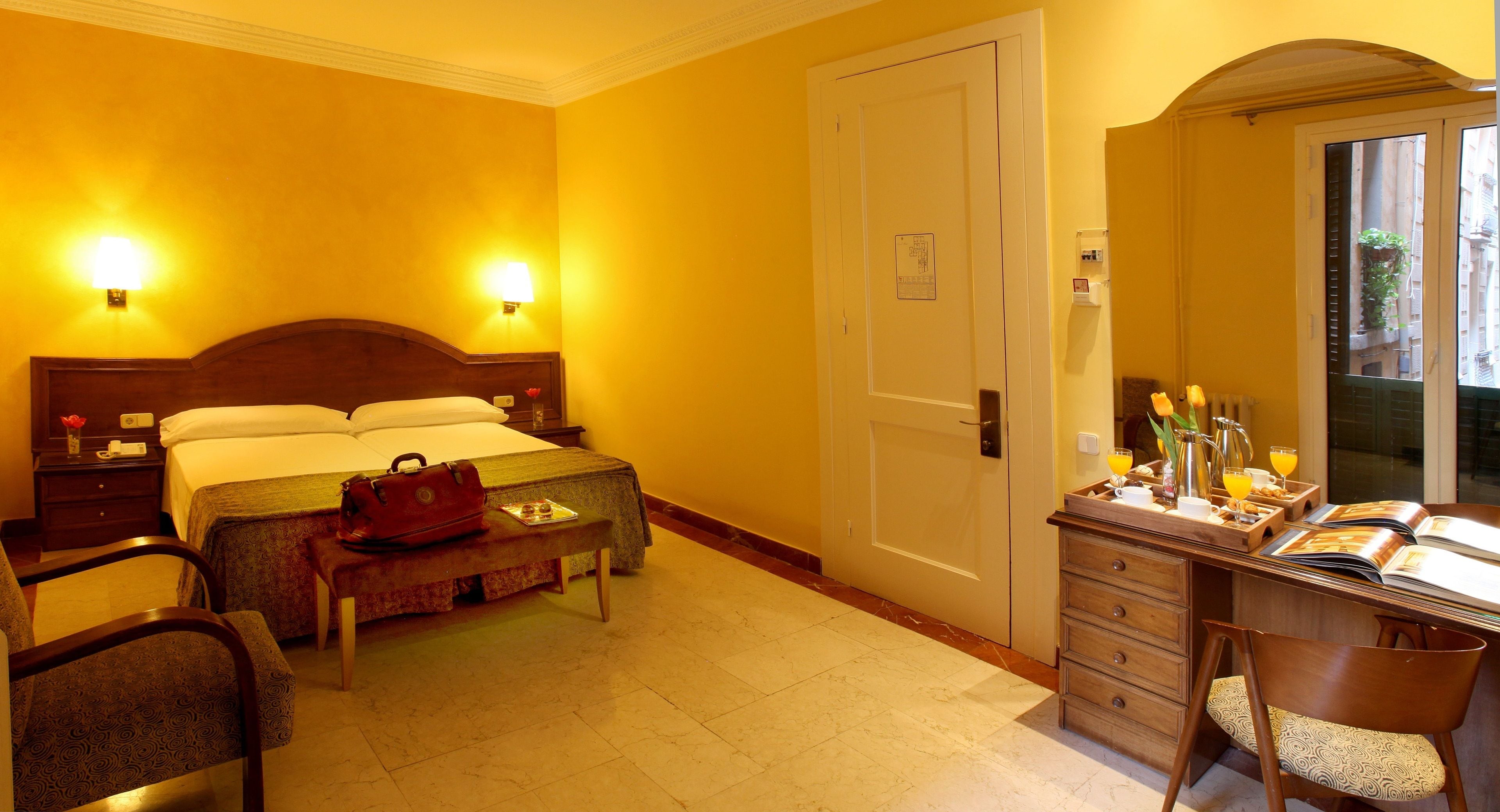 A hotel room with a bed, bedside tables, a desk with a chair, a bench, and a window with a view.