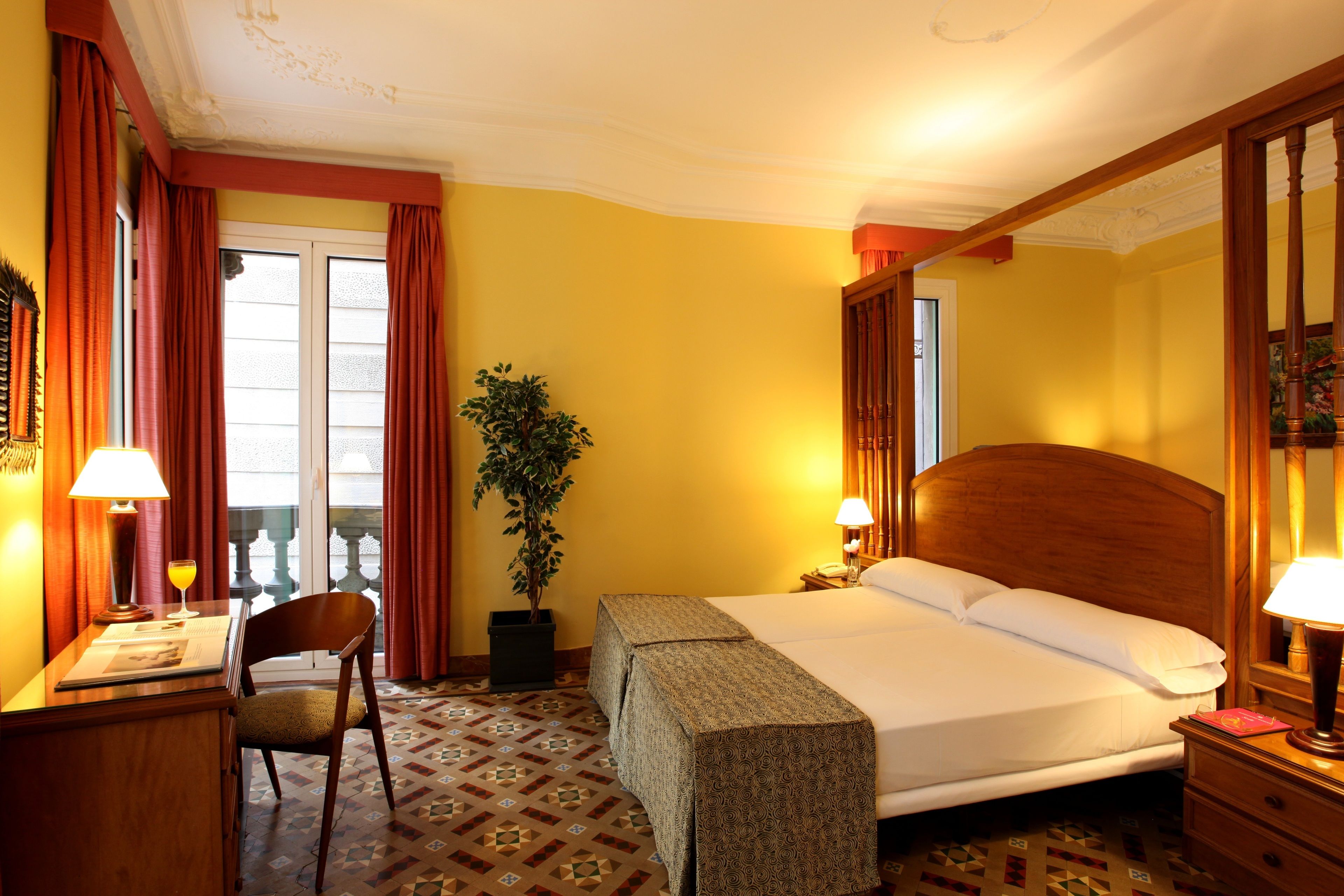 Ein Hotelzimmer mit Bett, Schreibtisch mit Lampe, Stuhl, Blumentopf und einem Fenster mit Vorhängen.