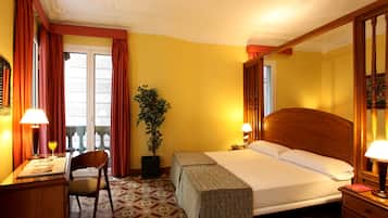 Habitación de hotel con cama, escritorio con lámpara, una silla, planta en maceta y ventana con cortinas.