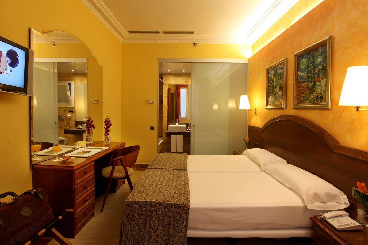 A hotel room with a bed, a desk with a chair, a dresser, a mirror, a TV, and artwork on the walls.