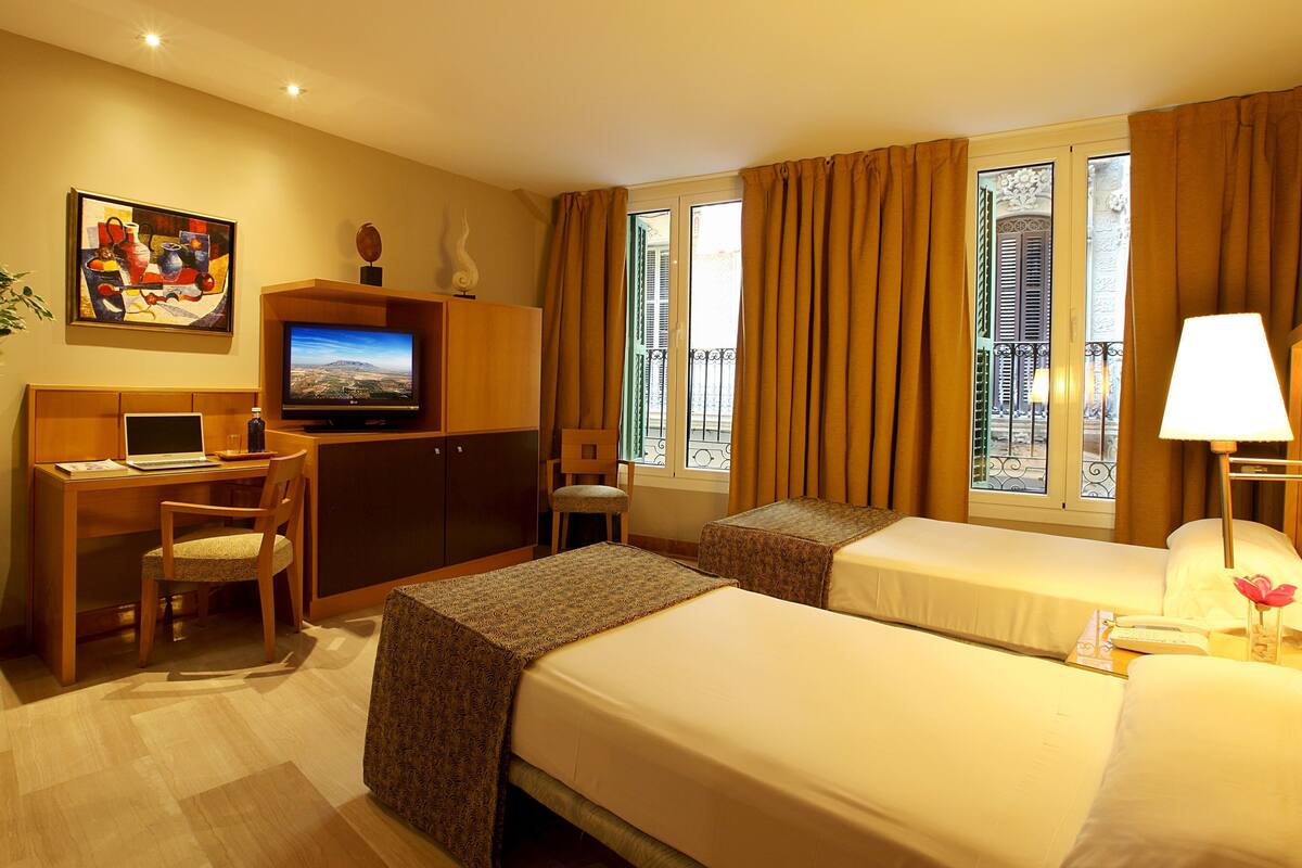 A hotel room with two beds, a desk with a laptop, a television, a potted plant, and a window with curtains.