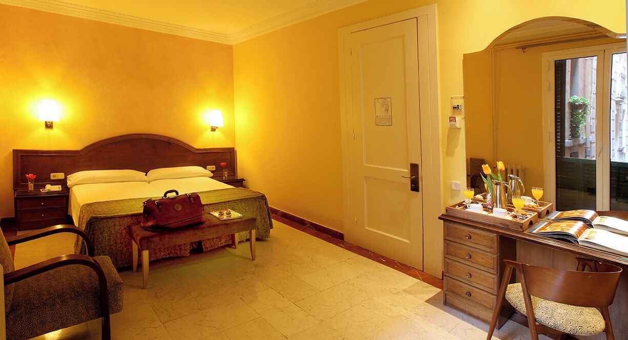 A hotel room with a bed, bedside tables, a desk with a chair, a mirror, and a window.