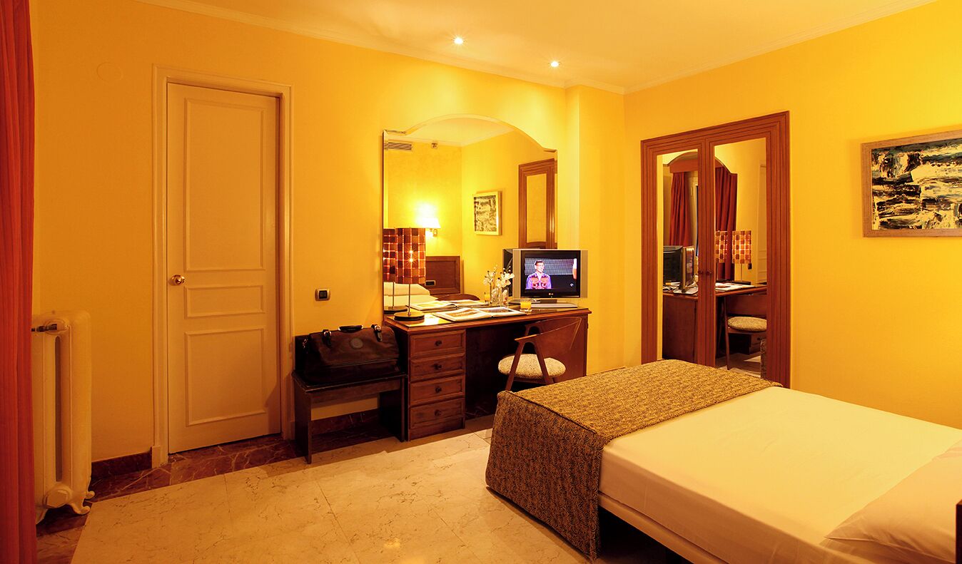 A hotel room with a bed, a desk with a computer, a chair, a radiator, and a door.