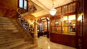 A grand staircase with ornate railings, a chandelier, and a wooden reception desk with glass panels. - Hotel Nouvel (Barcelona)