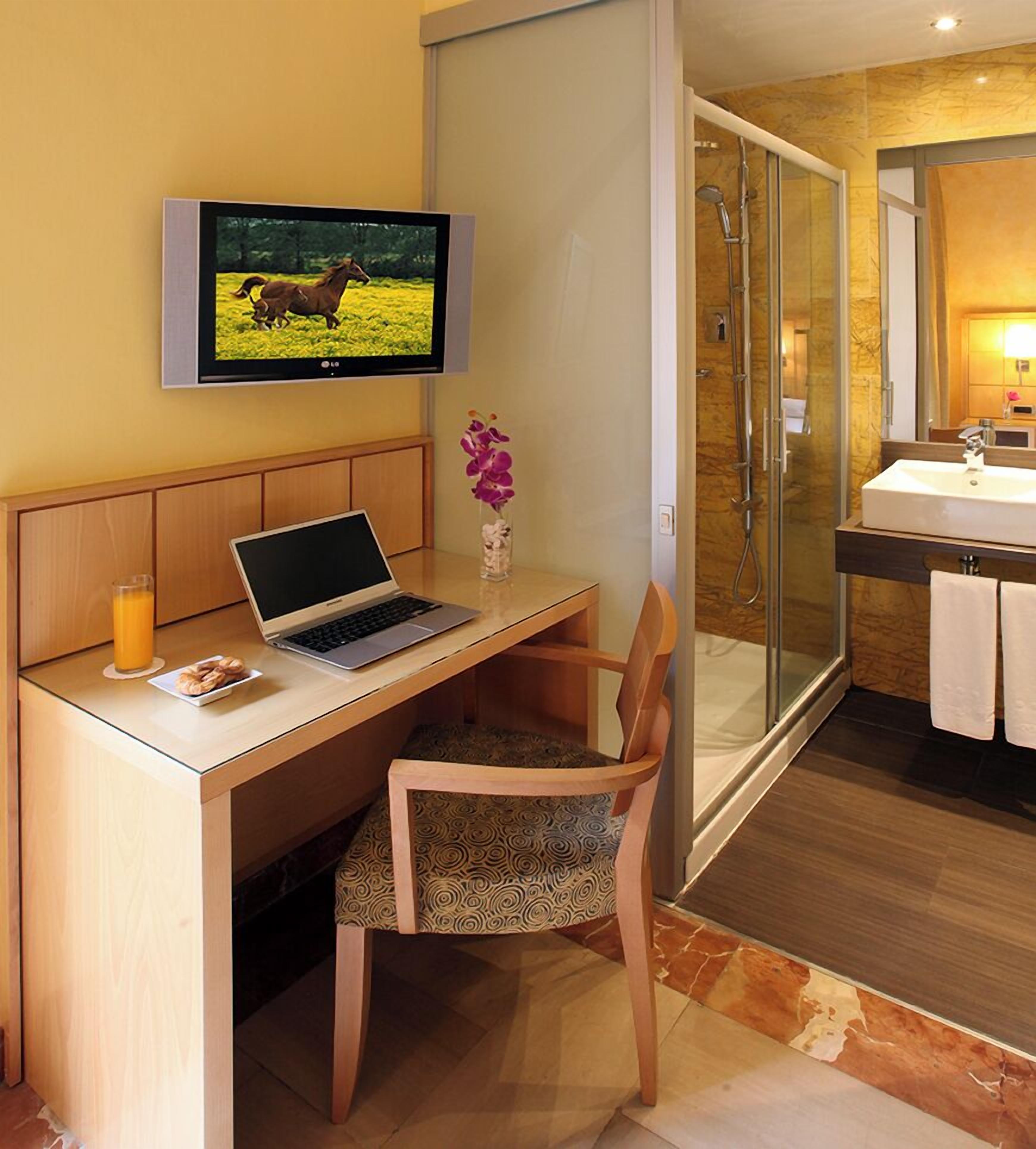 Habitación de hotel con un escritorio de madera, una computadora portátil, un televisor mostrando un caballo, un vaso de jugo de naranja y un área de ducha.