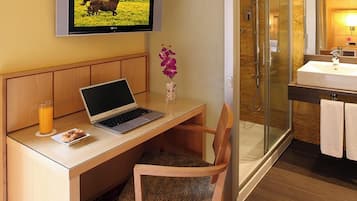 Habitación de hotel con un escritorio de madera, una computadora portátil, un televisor mostrando un caballo, un vaso de jugo de naranja y un área de ducha.