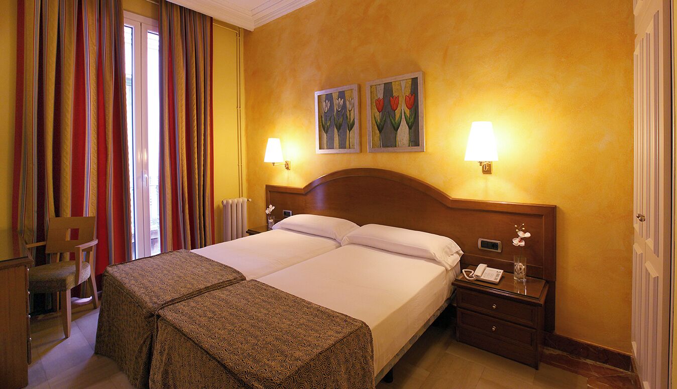 A hotel room with a double bed, two bedside tables, a chair, a desk, and a window with curtains.