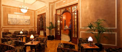 Un vestíbulo de hotel con sillas estampadas, mesas con lámparas, un candelabro y un cuadro grande en la pared.