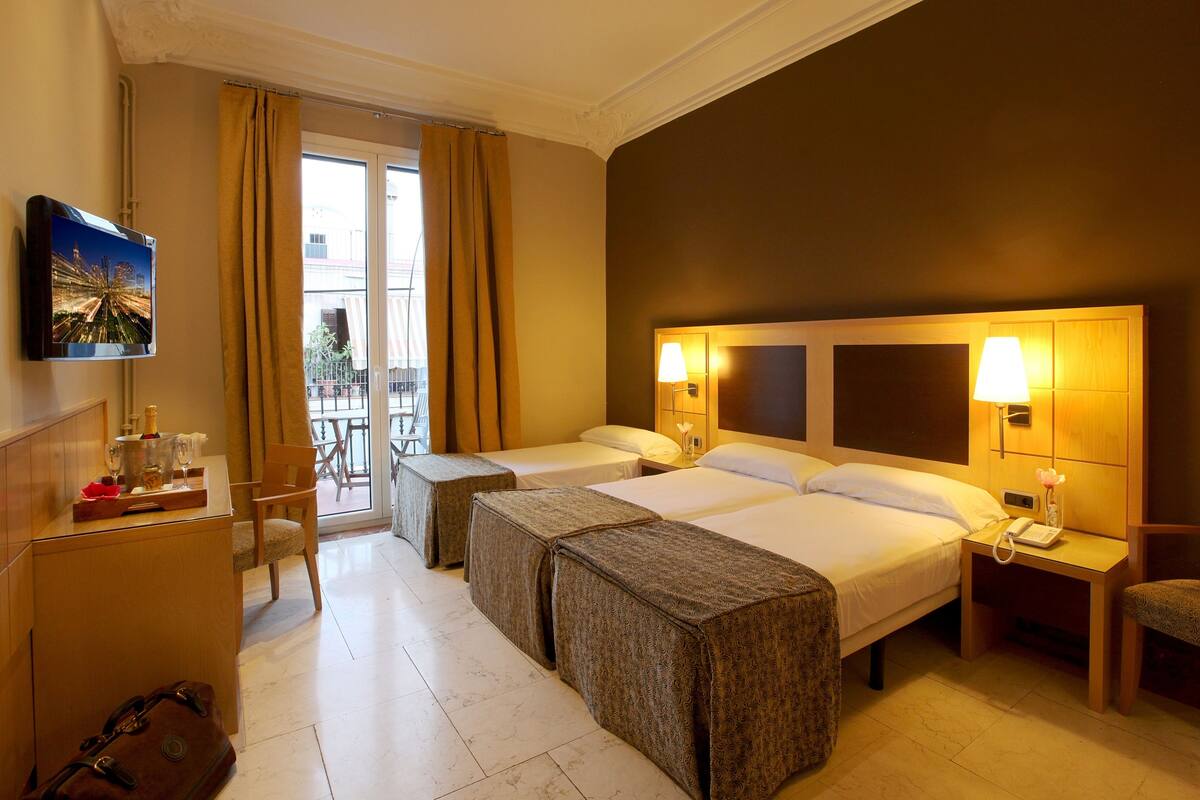 A hotel room with two beds, a desk with a TV, a chair, a small table with a bottle and glasses, and a view of the outside balcony.