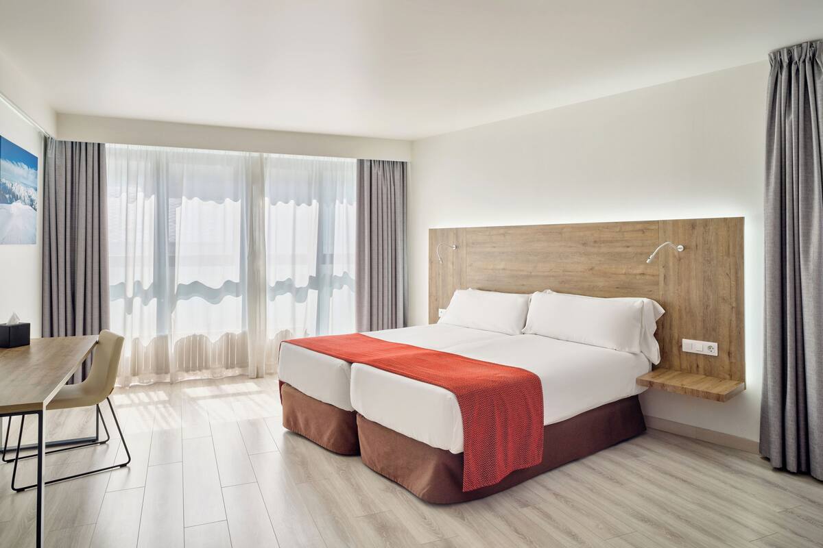 Ein modernes Hotelzimmer mit einem hölzernen Kopfteil, einem großen Bett mit roter Decke, einem Holztisch und Blick ins Freie durch transparente Vorhänge.
