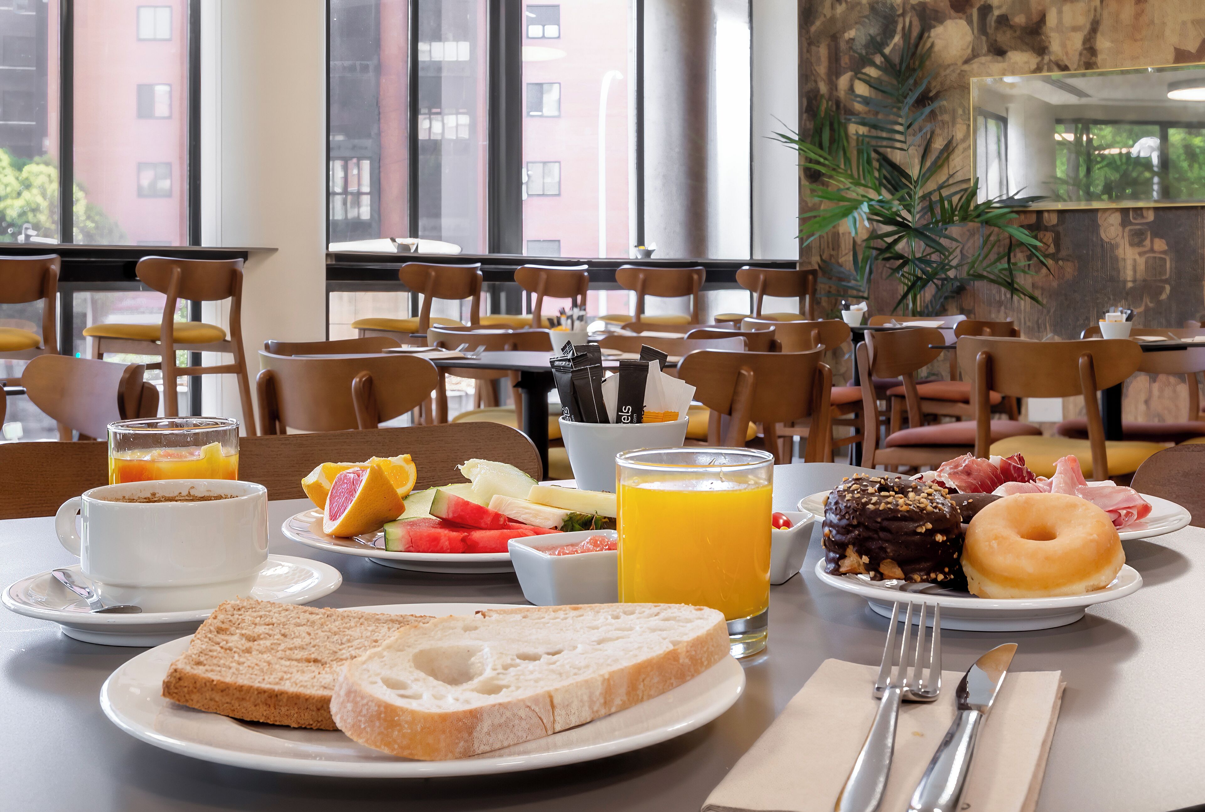Un desayuno con una mesa preparada para dos personas, que incluye tostadas, fruta, una rosquilla y un vaso de jugo de naranja.