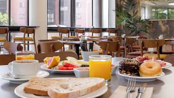 Un desayuno con una mesa preparada para dos personas, que incluye tostadas, fruta, una rosquilla y un vaso de jugo de naranja.