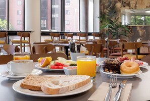 A breakfast setting with a table set for two, featuring plates of toast, fruit, a donut, and a glass of orange juice. - Hotel Acta Piramides (Madrid)