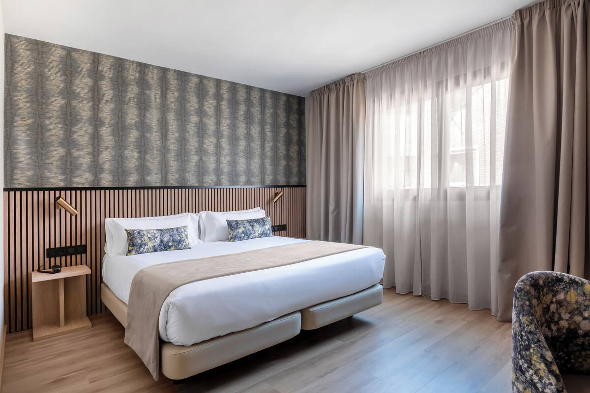 Una camera d'albergo moderna con un letto grande, un comodino, una sedia e una finestra con tende.