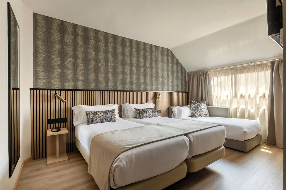 Una camera d'albergo moderna con due letti, una testiera in legno a listelli, un comodino e un'ampia finestra con tende.