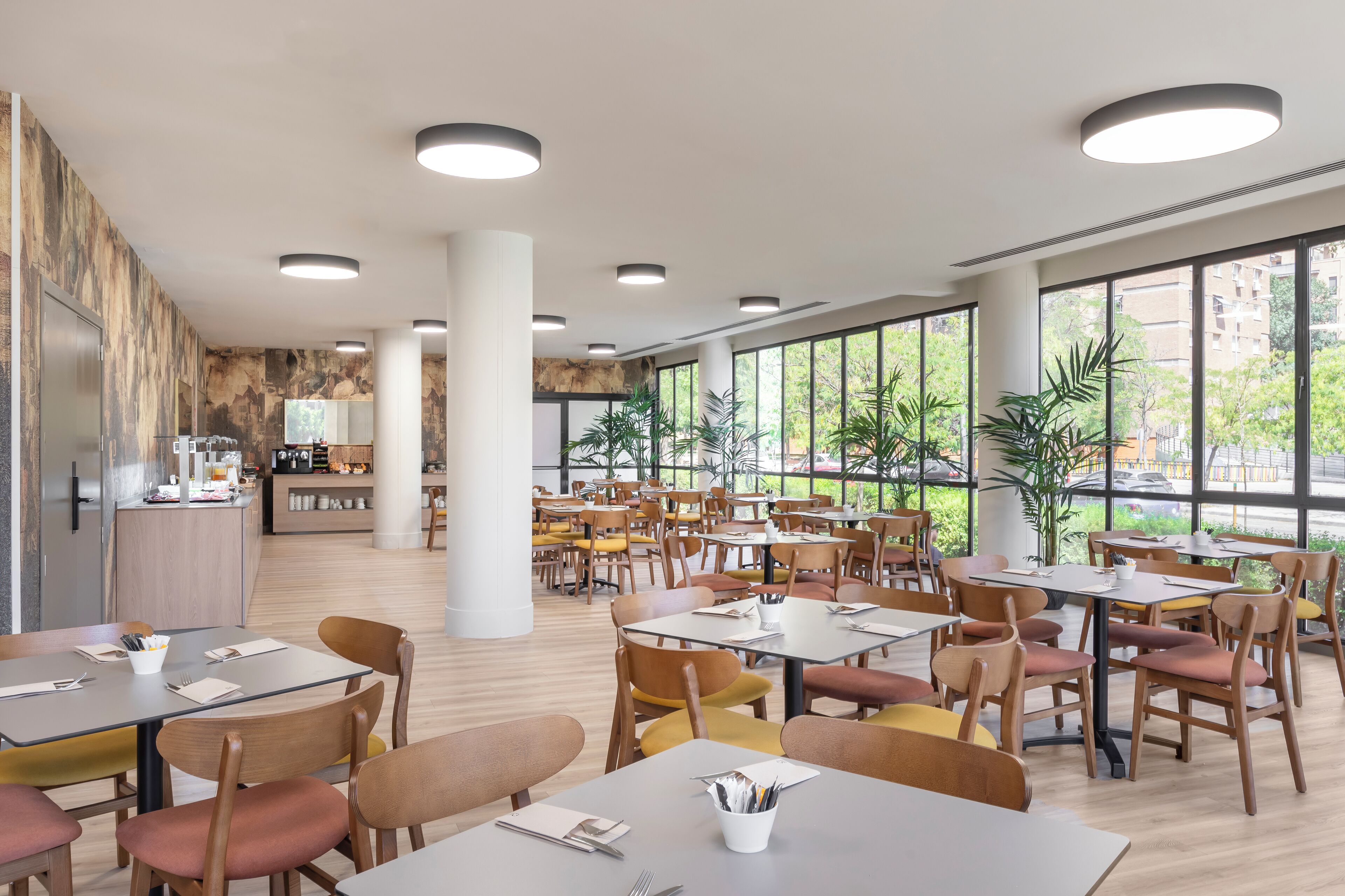 A spacious dining area with multiple tables and chairs, large windows offering views of greenery and buildings, and a kitchen area in the background.