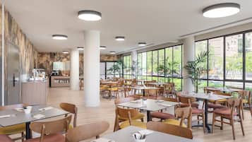 A spacious dining area with multiple tables and chairs, large windows offering views of greenery and buildings, and a kitchen area in the background.
