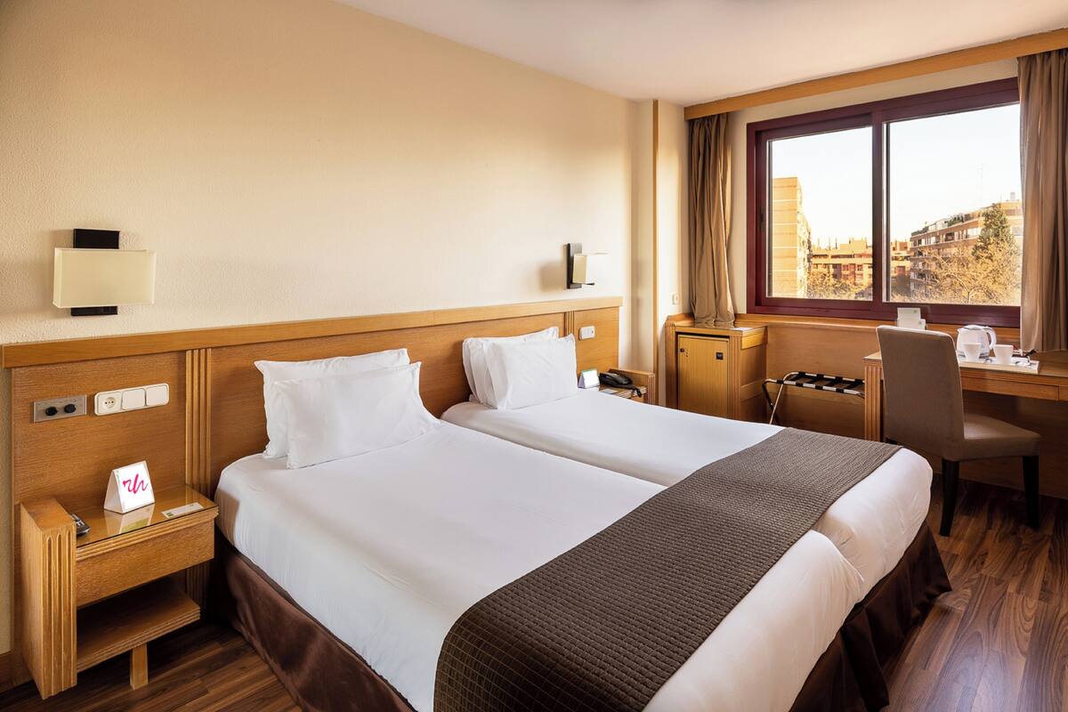 Une chambre d’hôtel avec deux lits, un bureau, une chaise, une table de chevet et une fenêtre donnant sur la ville.