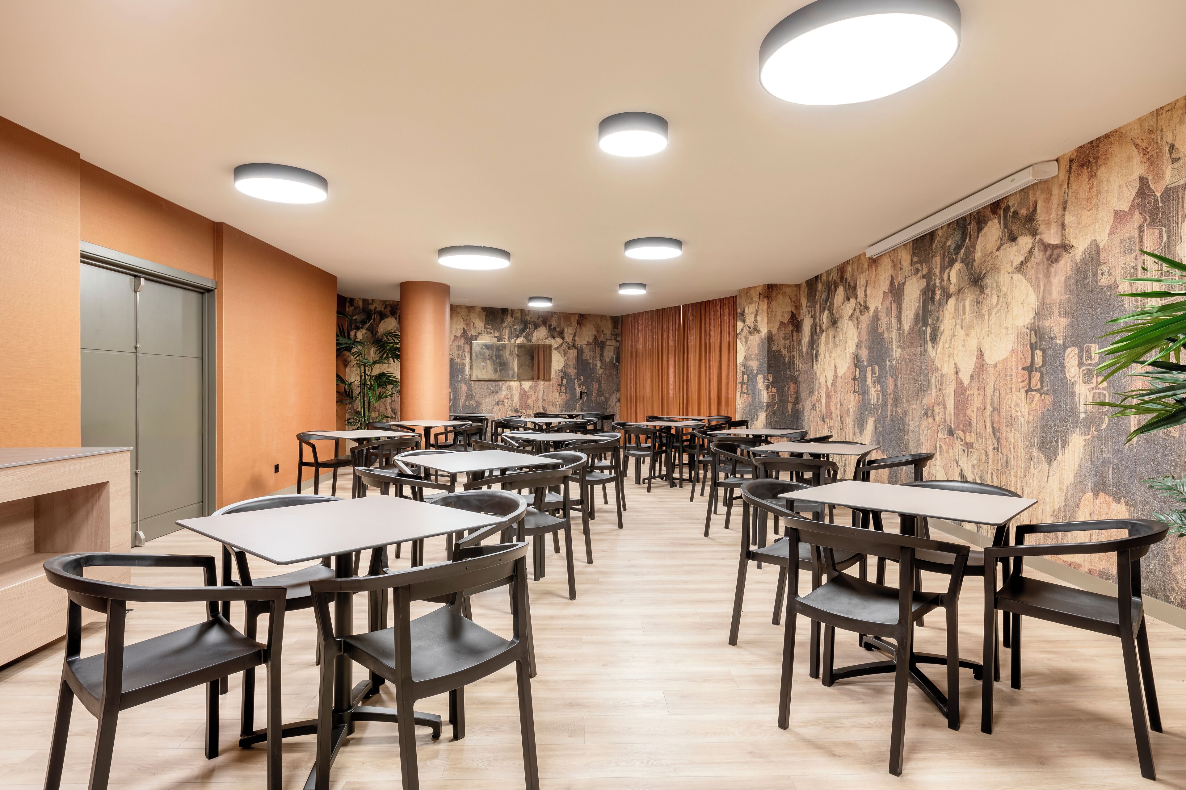 Un salón de conferencias con varias mesas y sillas, luces empotradas en el techo y un mural en la pared.