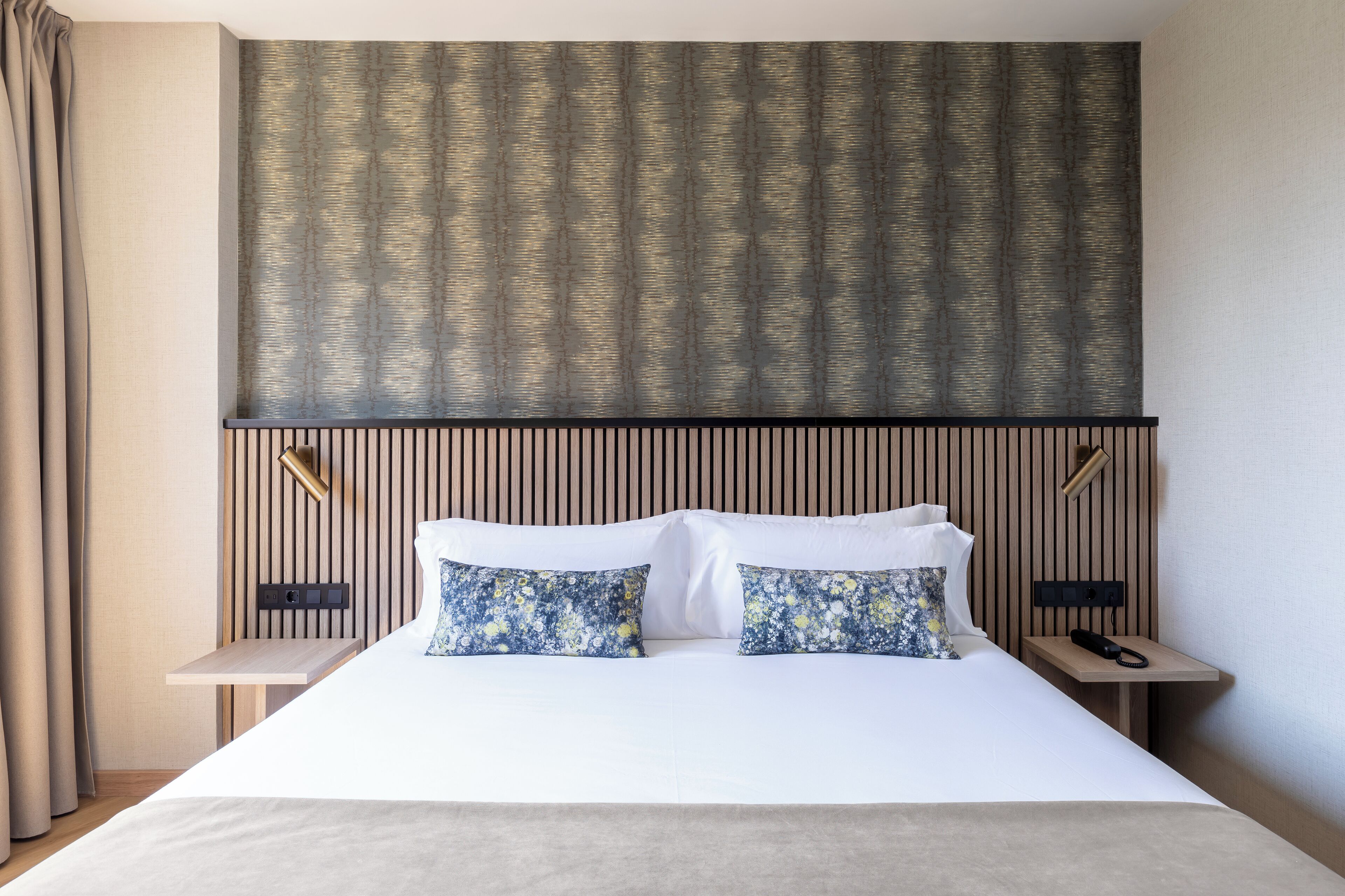 Una habitación de hotel moderna con una cama grande, dos mesitas de noche, un cabecero con líneas verticales y una pared con diseño.
