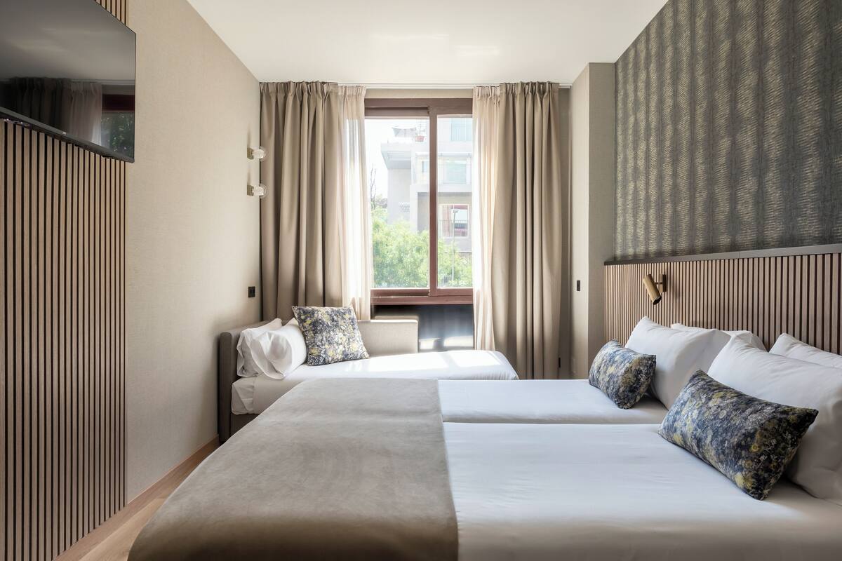 Camera d'albergo con due letti, TV a muro, finestra con tende e vista sugli edifici.