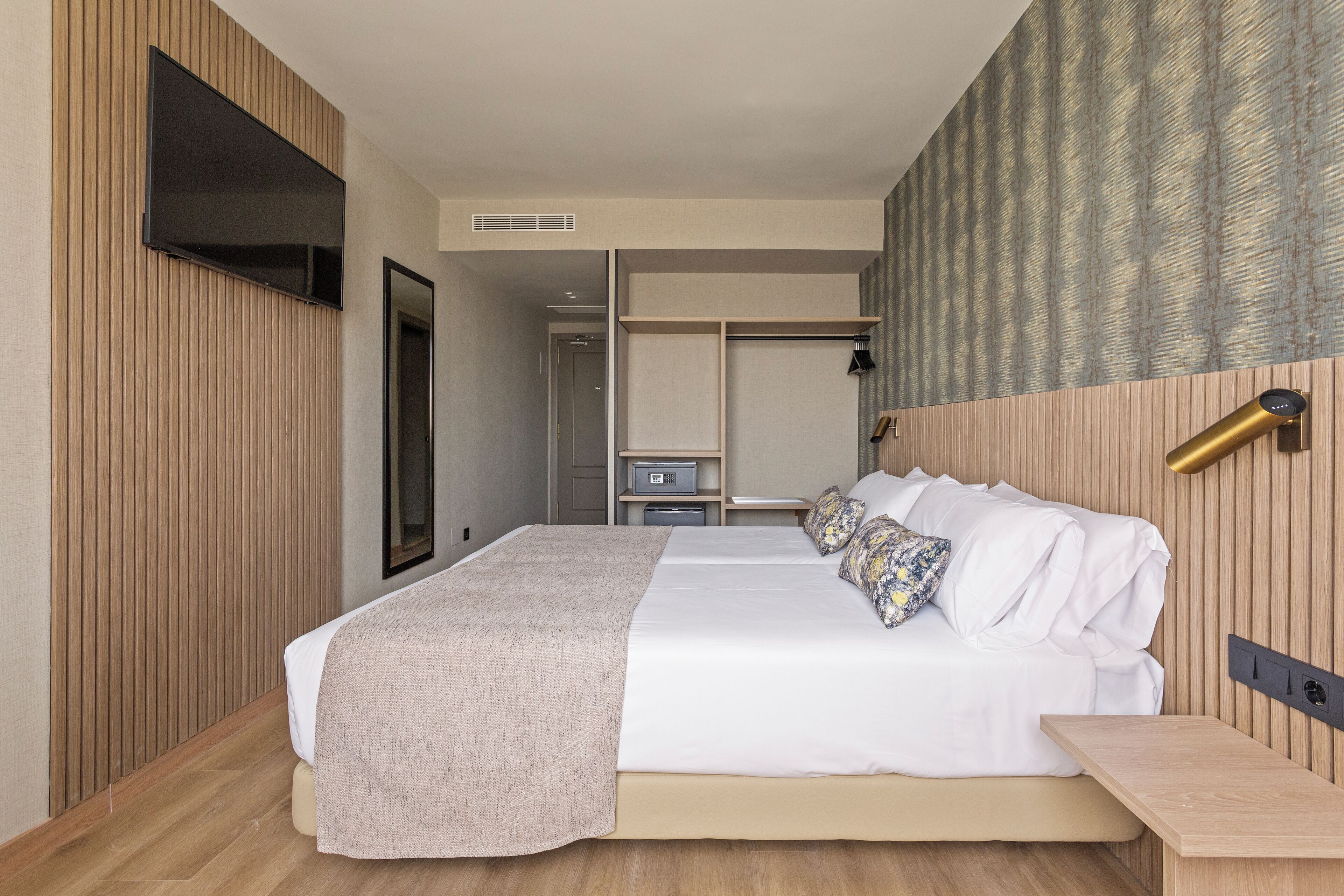 Una habitació d'hotel moderna amb un llit gran, una televisió penjada a la paret, un armari i una taula de nit amb una làmpada.