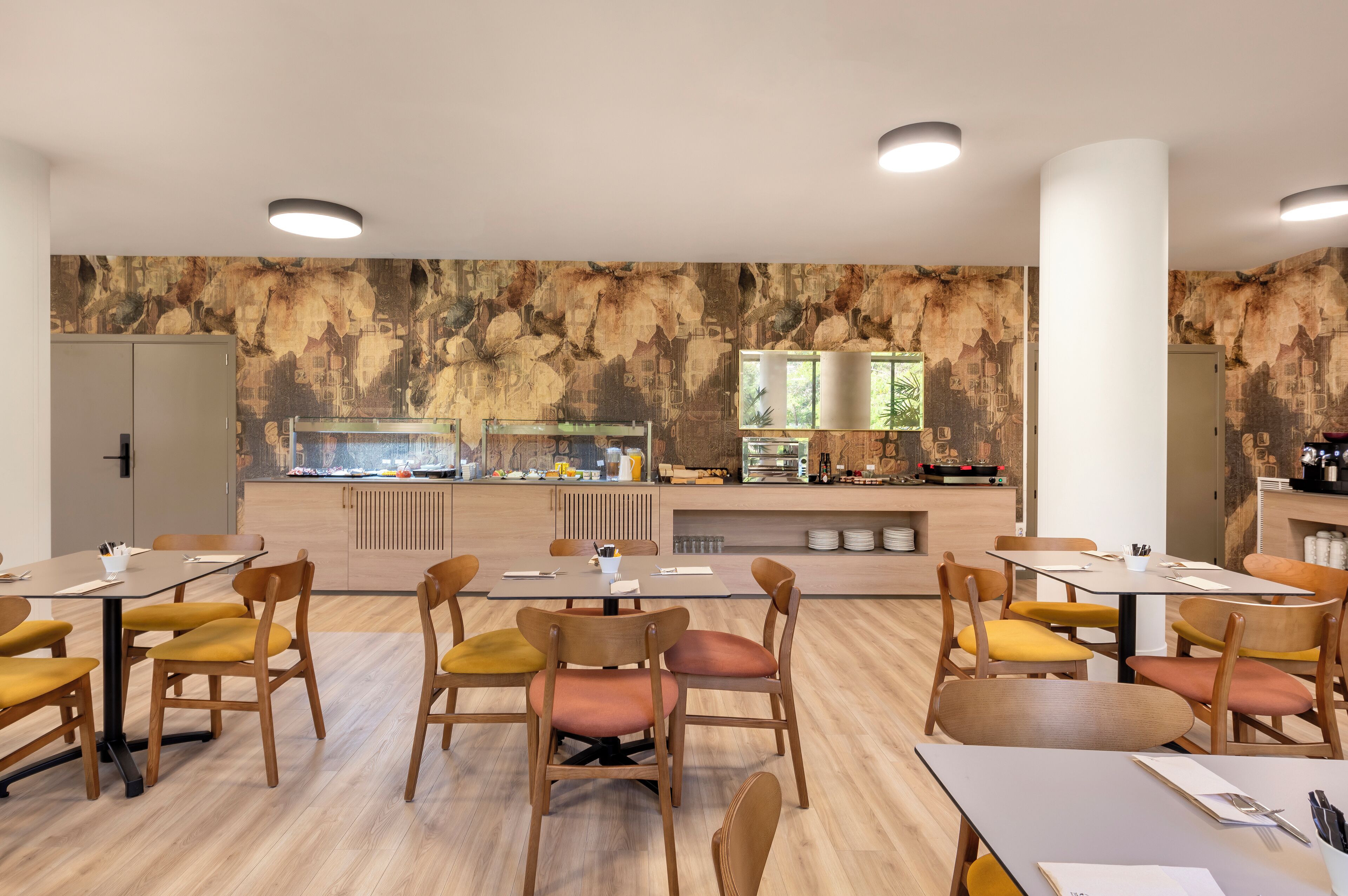 A modern dining area with wooden tables and chairs, a kitchen counter with food display, and a stone wall backdrop.