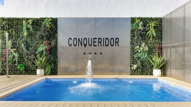 A swimming pool with a fountain, surrounded by lush greenery and a wall with the word 'CONQUERADOR' and stars. - Hotel Conqueridor (Valencia)