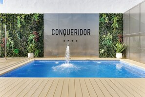 A swimming pool with a fountain, surrounded by lush greenery and a wall with the word 'CONQUERADOR' and stars.