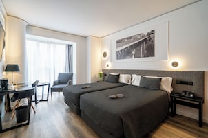 Habitación de hotel con dos camas, un escritorio, una silla, una mesita de noche y un cuadro en la pared.