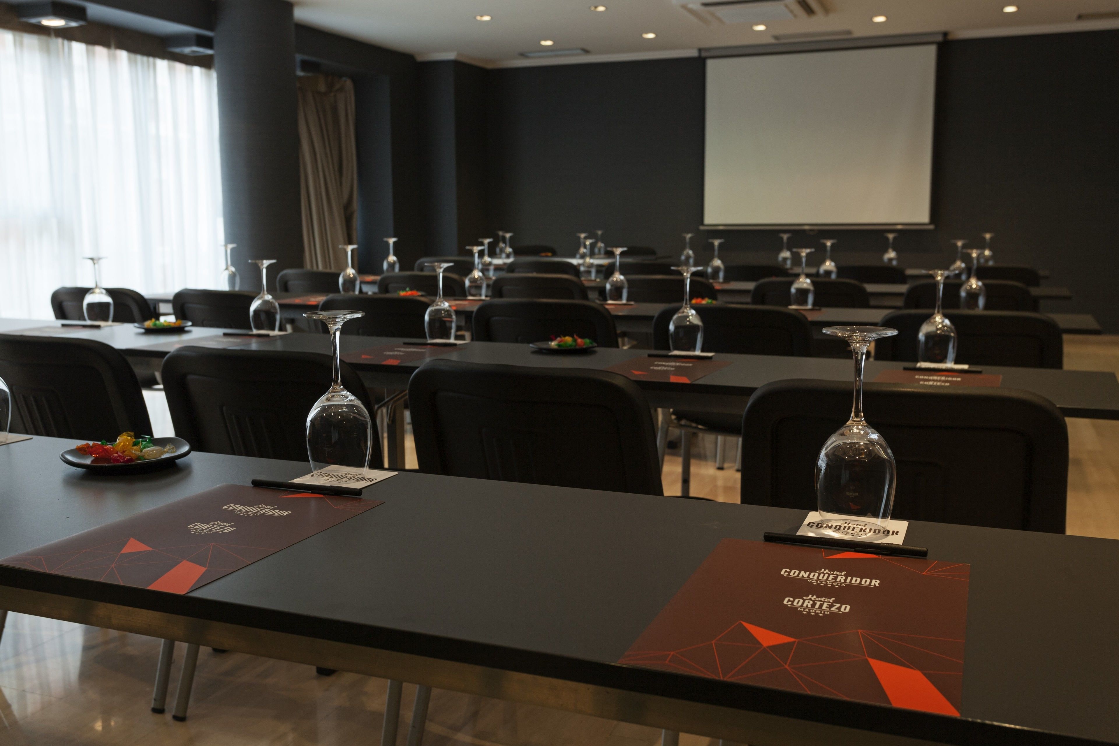 Ein Konferenzraum mit Tischen, die für eine Besprechung gedeckt sind, jeweils mit Namensschild, Weingläsern und einer kleinen Fruchtschale.