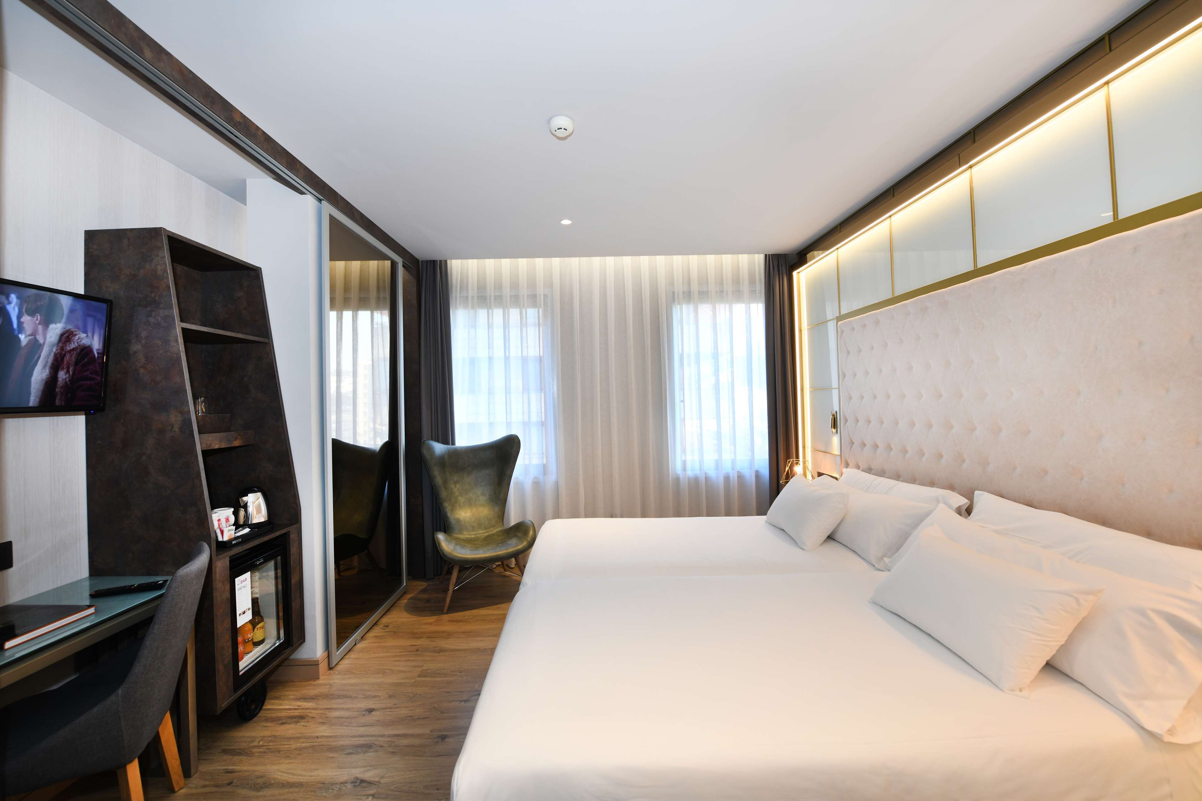 Una habitación de hotel moderna con una cama grande, un escritorio con computadora, un televisor y una silla.
