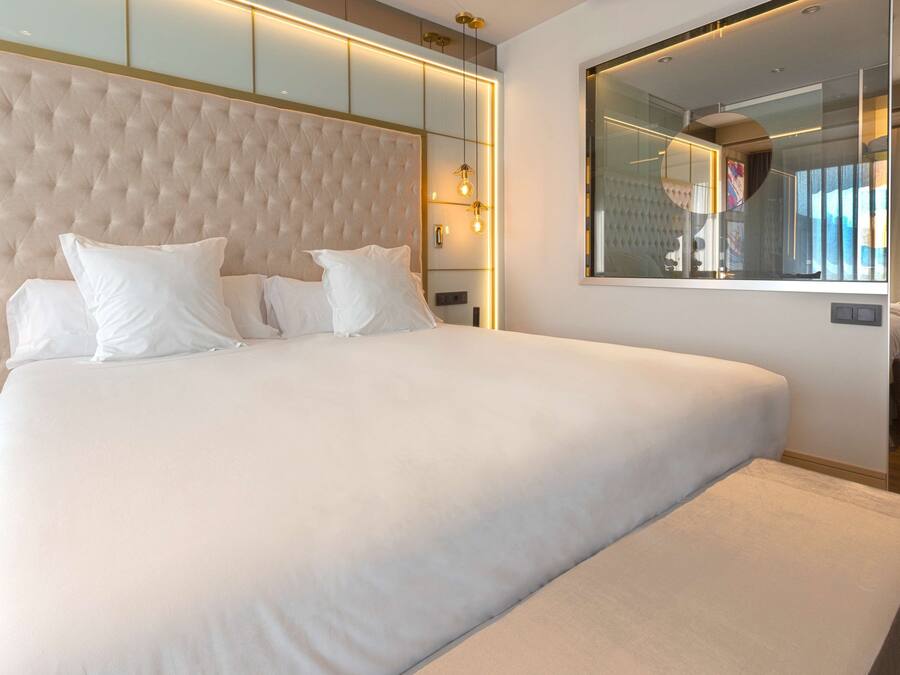 Una cama grande con cabecera capitonada, una mesita de noche y un espejo fijado a la pared.