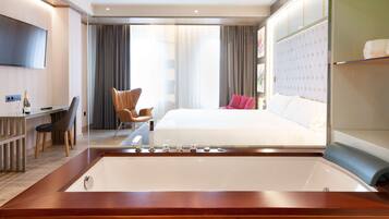 A modern hotel room with a large bed, a desk with a chair, a television, and a bathtub.