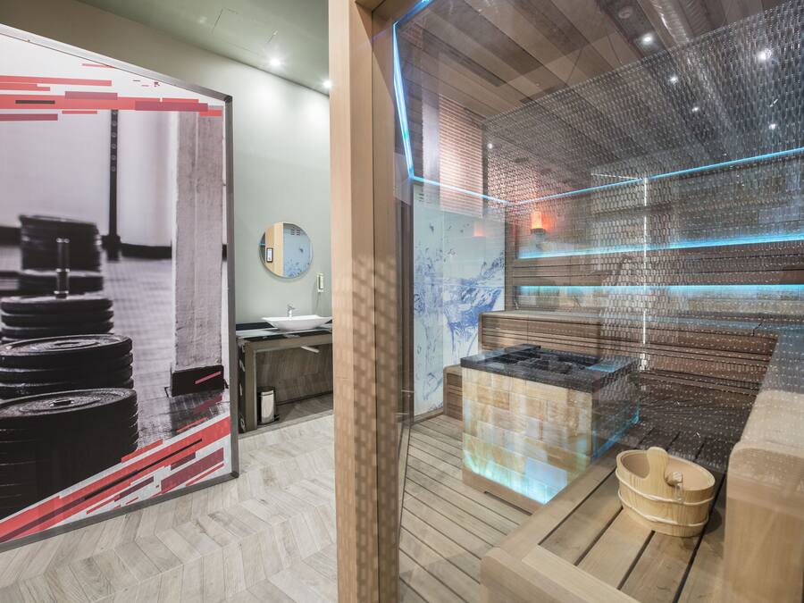 Un espace spa moderne comprenant un sauna, des bancs en bois et une paroi en verre donnant sur la salle de sport.