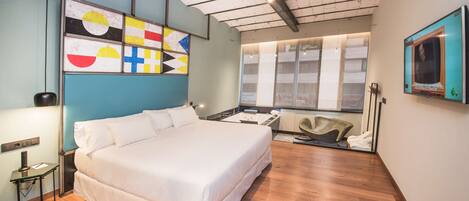 A modern hotel room with a large bed, a desk, a chair, a TV, and a window with blinds.