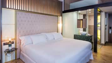 A modern hotel room with a large bed, a bedside table with a lamp, a bathroom visible through an open door, and a wooden slatted partition above the bed.