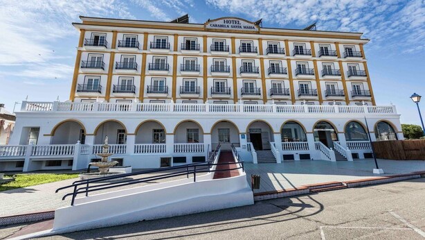 A multi-story hotel with a white facade and yellow trim, featuring balconies and a central courtyard with a fountain. - Hotel Carabela Santa Maria (Moguer)