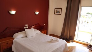 A hotel room with a bed, a nightstand, a wall-mounted light, a painting, and a window with curtains. - Hotel Carabela Santa Maria (Moguer)