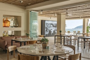 A spacious dining area with round tables, comfortable seating, and a large window offering a view of the sea.