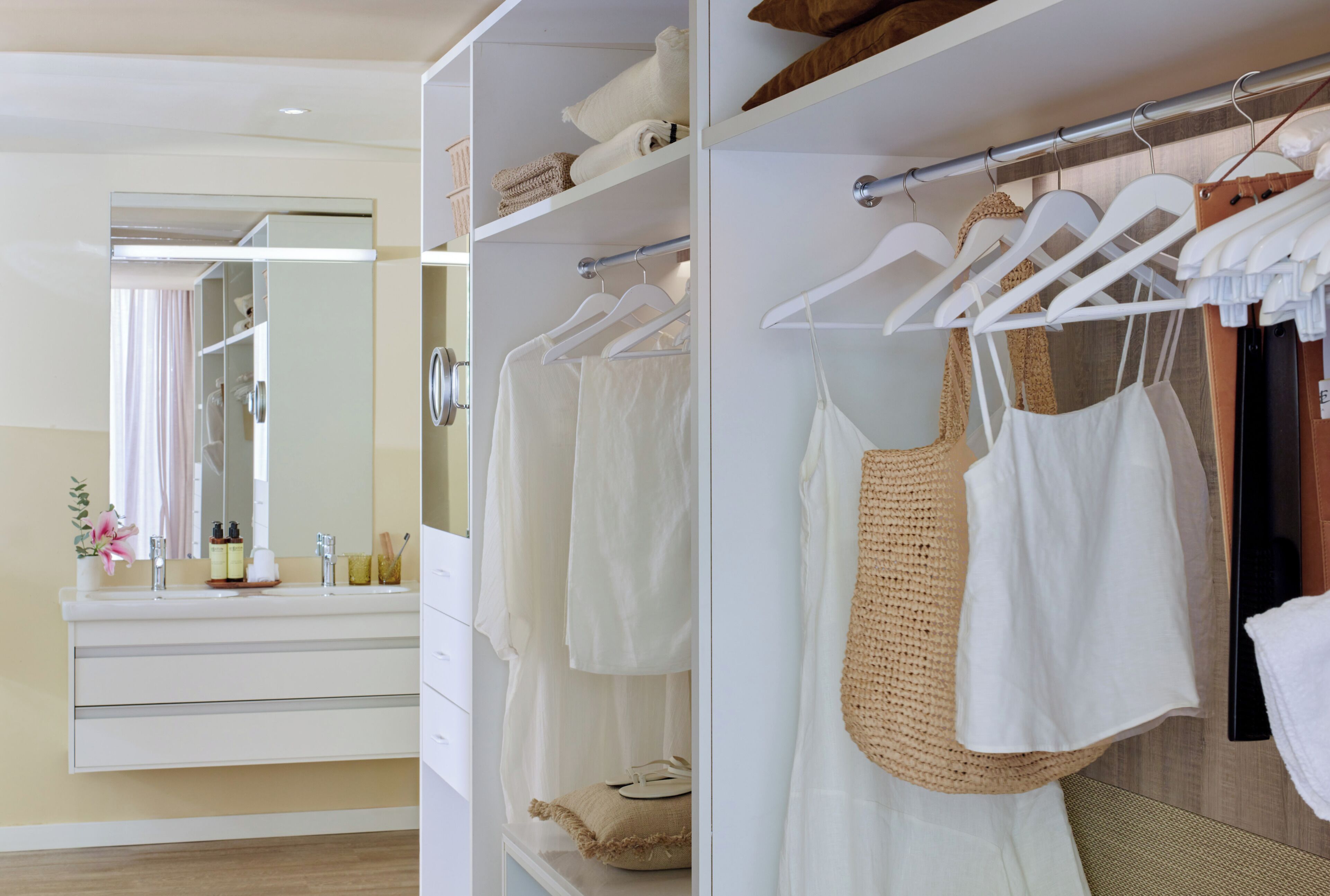 A hotel room with a large mirror, a vanity with toiletries, a wardrobe with hanging clothes, and a bed with a white comforter.