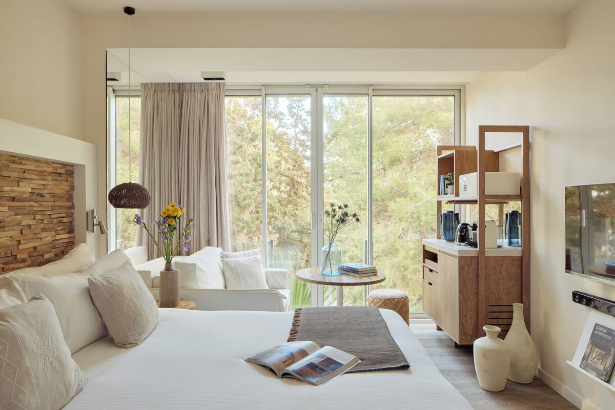 A modern bedroom with a large bed, a wooden headboard, a small table with books and flowers, a wooden cabinet, and a large window with a view of trees.