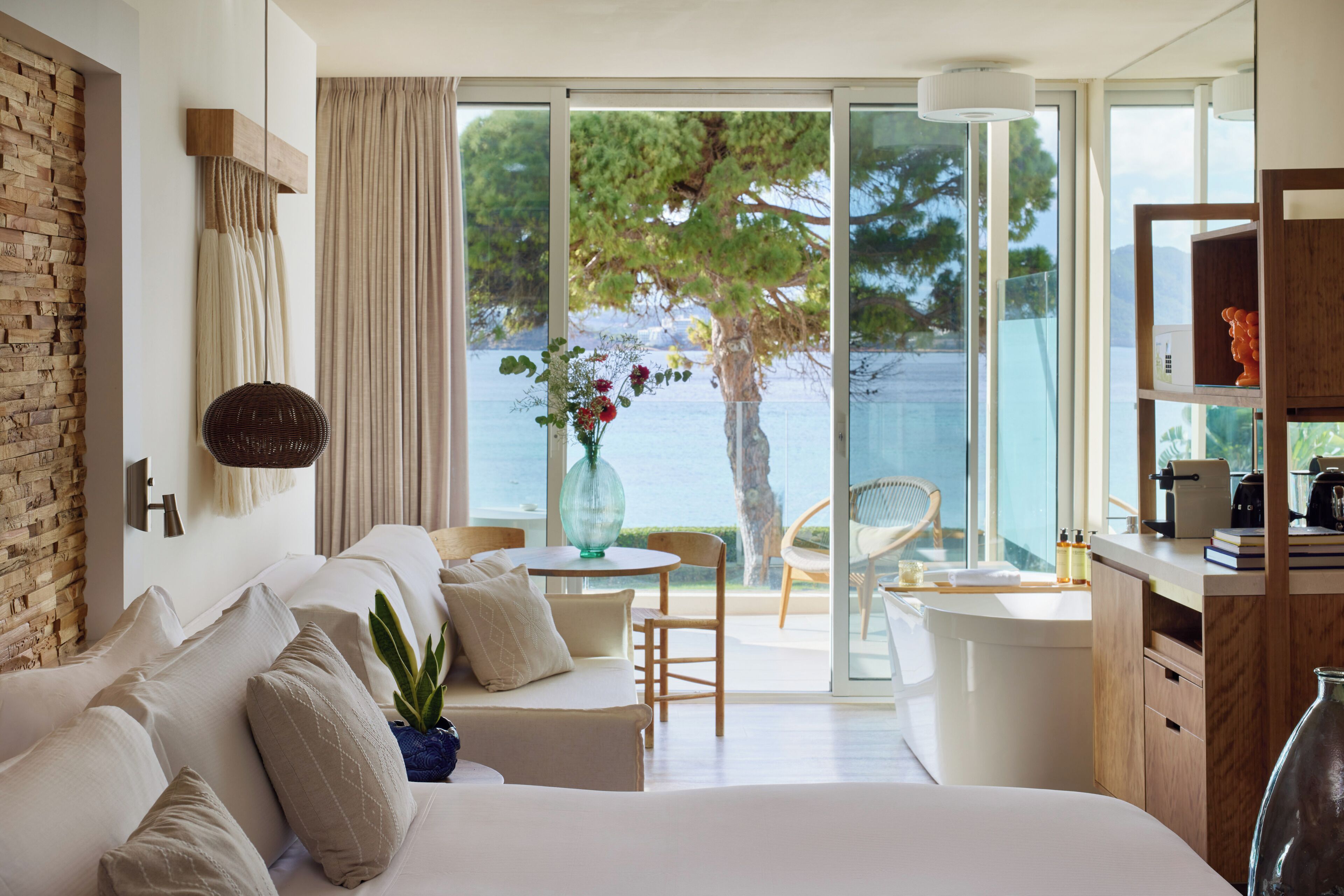 Ein modernes Hotelzimmer mit einem großen Bett, einer Sitzecke mit Tisch und Stühlen, Meerblick und einem Balkon mit einem Sessel.