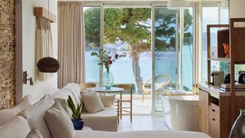 Ein modernes Hotelzimmer mit einem großen Bett, einer Sitzecke mit Tisch und Stühlen, Meerblick und einem Balkon mit einem Sessel.