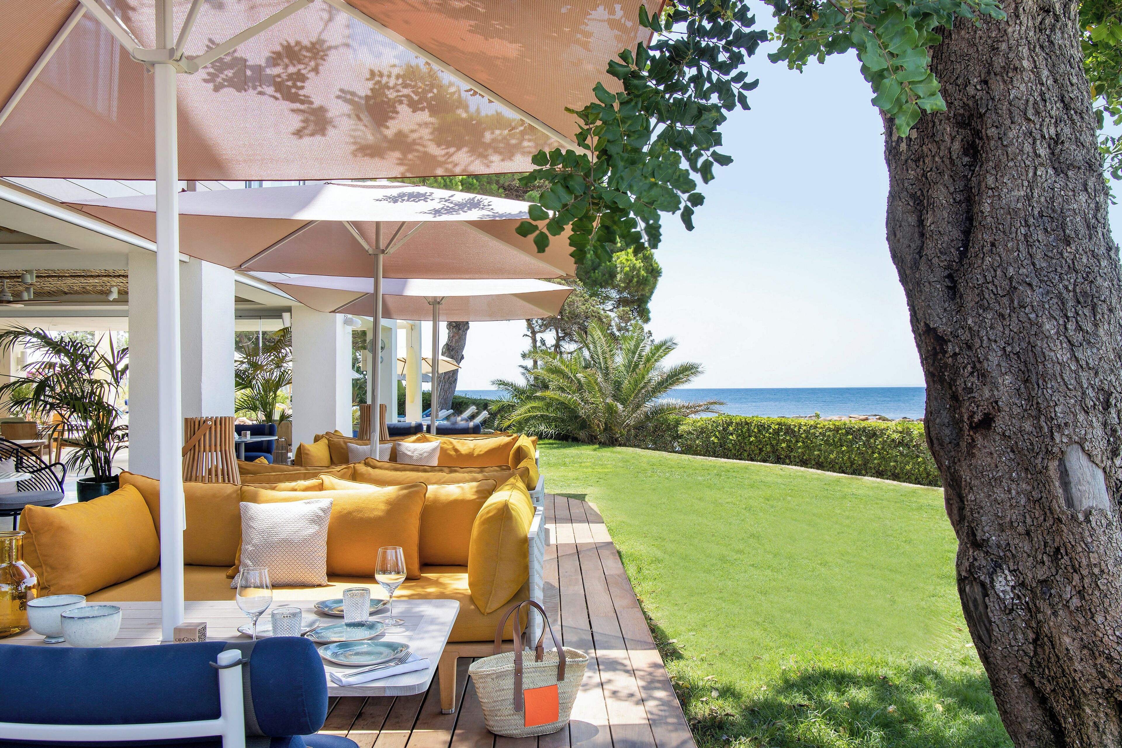 A sunlit outdoor seating area with a view of the sea, featuring a wooden deck, yellow cushions, and a table set with glasses and a basket.